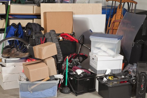 A collection of various household items and rubbish placed together in an indoor storage or garage area. Visible objects include cardboard boxes, plastic storage containers, a black toolbox, a partially visible bicycle wheel, and miscellaneous items such as paper, fabric, and small tools. The items are stacked and arranged haphazardly on a concrete floor with a background featuring shelving units and furniture pieces, indicating an ongoing clearance or rubbish removal process. The scene showcases clutter typical of household rubbish that a company like Office Clearance Lee might assist with in the Lee area or postcode region. The lighting appears natural, and the environment is typical for a domestic storage space awaiting rubbish collection or disposal. Overall, the image illustrates the type of clutter targeted by commercial and residential rubbish removal services, with a focus on general household rubbish and waste awaiting clearance.