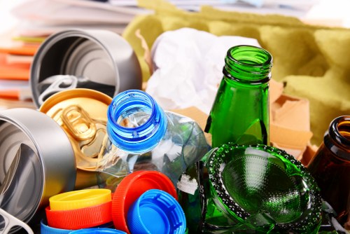 A collection of discarded glass bottles, including green and brown varieties, along with plastic bottles featuring blue and yellow caps, all weathered and piled together on a surface, with some lids and caps scattered nearby. In the background, partially visible, are pieces of crumpled paper or packaging and a yellow waste sack, suggesting a waste collection or rubbish removal scenario typical of commercial office clearance services. The sunlight highlights the reflective textures of the glass bottles and plastic caps, emphasizing their cleanliness and the collection's informal nature. The scene is set outdoors or in a warehouse environment, aligning with typical rubbish disposal or clearance activities in the Lee area. Office Clearance Lee often handles similar waste removal tasks involving recycling and proper disposal of mixed waste materials, particularly bottles and plastics, in line with environmental standards in the UK.