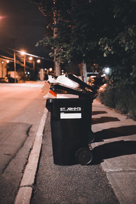 A person wearing a grey and yellow checkered jacket, a green long-sleeve shirt, and grey trousers, is holding open a large black plastic rubbish bag with light green gloves on both hands. The bag is p
